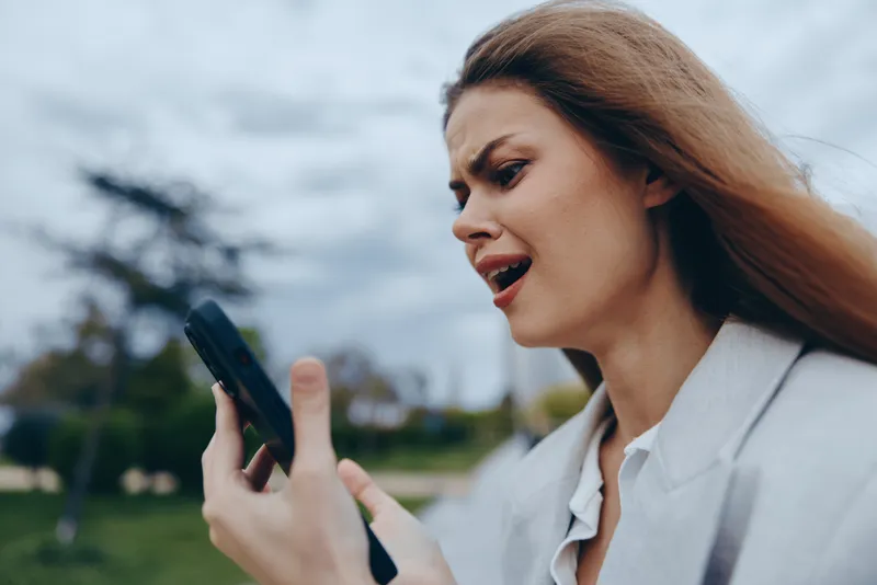 Woman experiencing frustration with smartphone outdoors in casual attire, emotional expression, cloudy sky background, autumn season concept