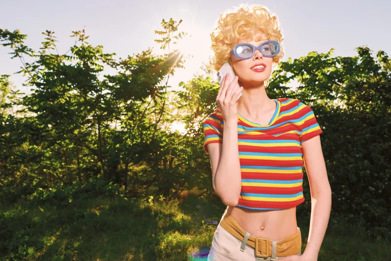 Smiling woman with curly blonde hair wearing a striped crop top and sunglasses. Outdoor scene with warm sunlight, retro vibe, and casual lifestyle. Colorful, nostalgic mood.
