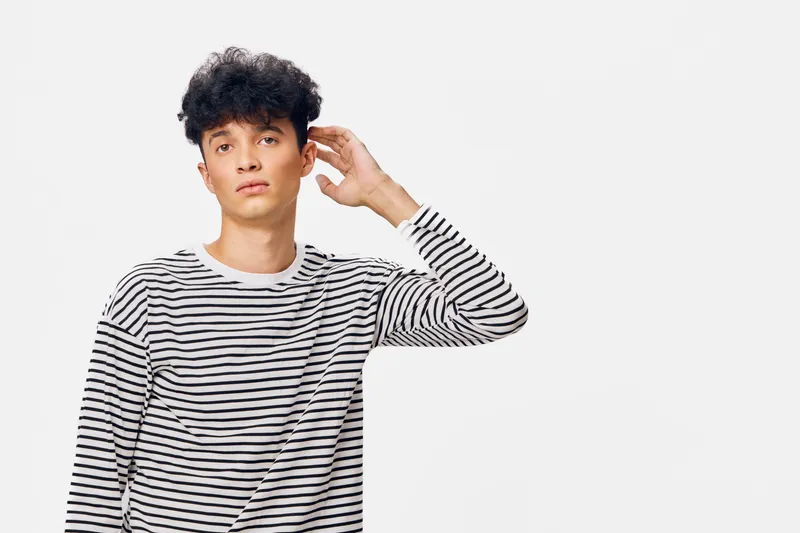 young man wearing striped black and white long sleeve shirt touching ear with hand, standing on white background with thoughtful expression