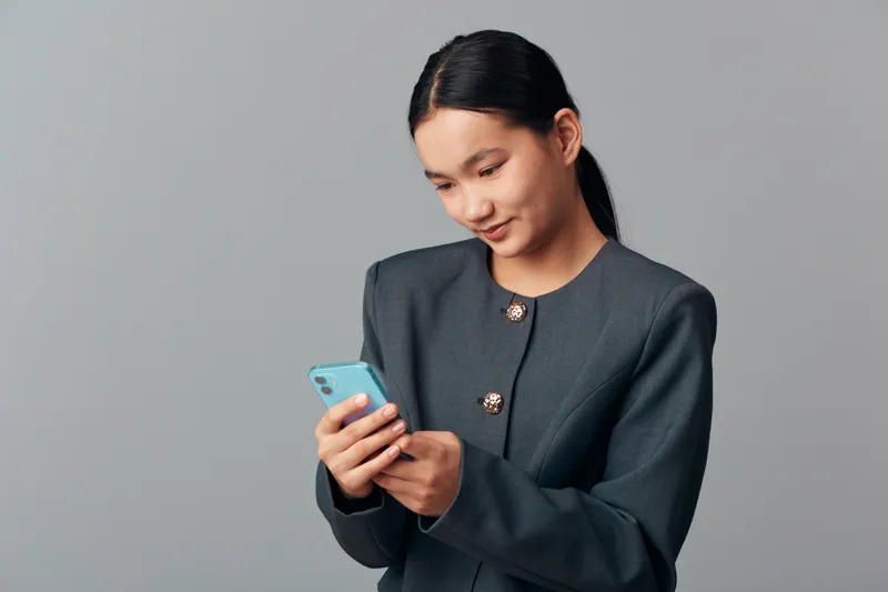 Young woman in dark jacket holding smartphone while smiling and looking at screen standing against grey background in studio. Technology, communication, casual and portrait concept.