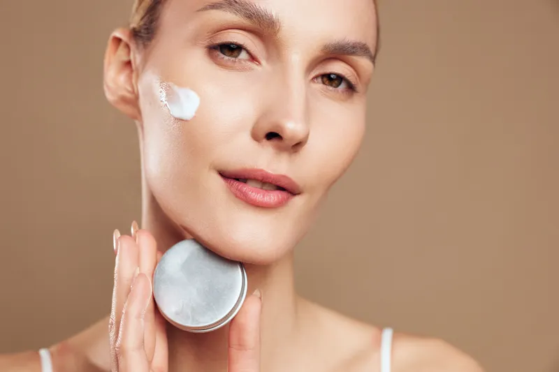 Young woman applies face cream on cheek holds cosmetic jar close to skin demonstrates skincare routine with natural makeup and smooth complexion on beige background.