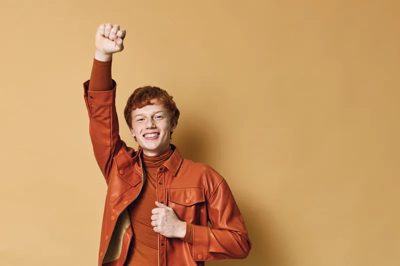 Young man with red hair wearing orange jacket and turtleneck sweater raising fist in air and smiling against beige background for success and confidence.