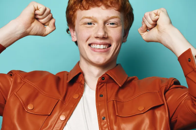Young man with red hair smiling and showing fists in brown jacket against blue background. Portrait of excited happy person with cheerful expression and raised hands.