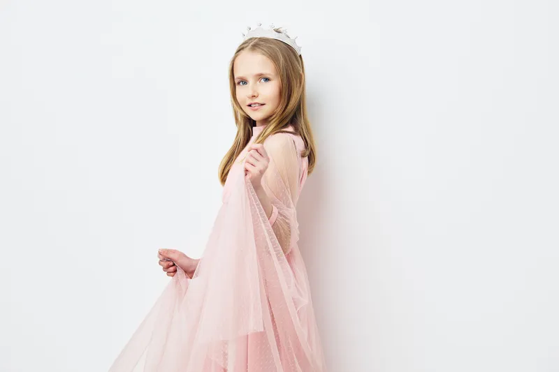 Young girl in a pink dress posing against a white background, showcasing innocence and playfulness with a tiara, perfect for children s fashion themes