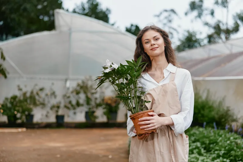 Young woman in a greenhouse holding a potted plant, wearing a light outfit and smiling, surrounded by greenery, representing gardening and sustainable living