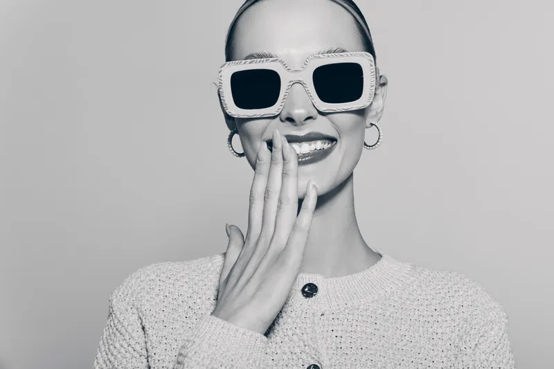 Smiling white woman with pale skin wears oversized sunglasses and knitted sweater, covering mouth with hand. Concept of fashion and joyful mood.