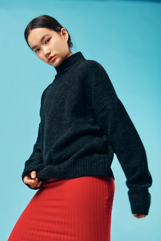 Young woman wearing black sweater and red skirt posing against blue background in studio. Fashion portrait, casual style, confident female model with minimal makeup and natural hair.