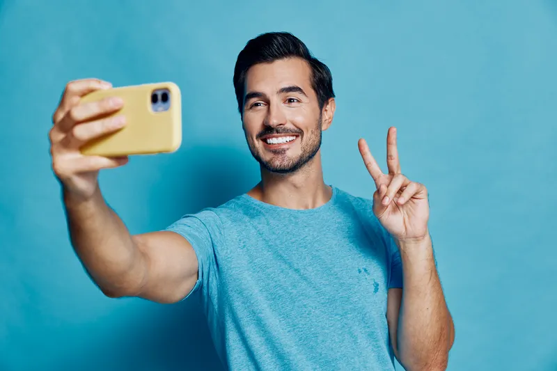Smiling man wearing a blue t-shirt taking a selfie with a smartphone, showing peace sign, isolated against a vibrant blue background. People lifestyle concept.