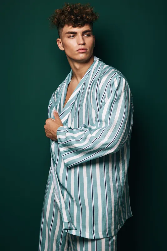 Young man with curly hair wearing striped pajamas standing against dark green background looking sideways with serious expression and hand on chest.