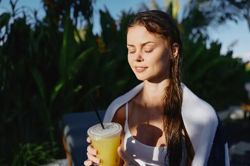 Cheerful woman outdoors, relaxed with a towel over her shoulders, holding a bright drink, sunlit moment, eyes closed, gentle breeze, natural colors, lush greenery background, casual summer lifestyle