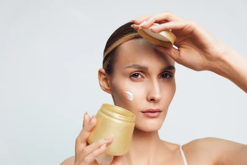 Woman applying moisturizer from a jar, facial cream on cheek, soft studio lighting, calm expression, skincare routine, neutral background, fresh natural tones, relaxed posture, soft shadows