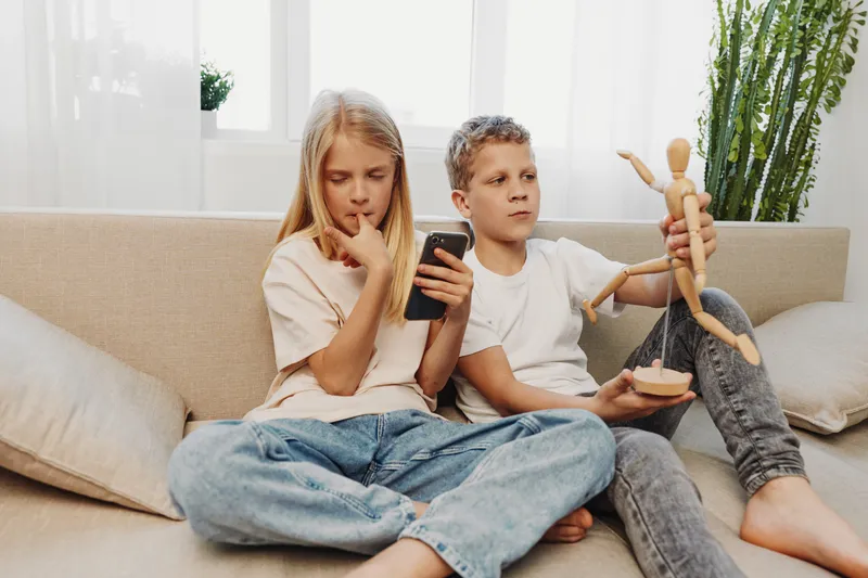Two children sitting on sofa at home, girl holding smartphone and boy playing with wooden mannequin, casual leisure moment indoors.