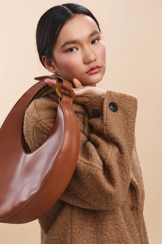 asian woman wearing a warm brown coat and carrying a curved leather bag poses against a soft neutral backdrop. calm and stylish, she exudes confidence and fashion sense.