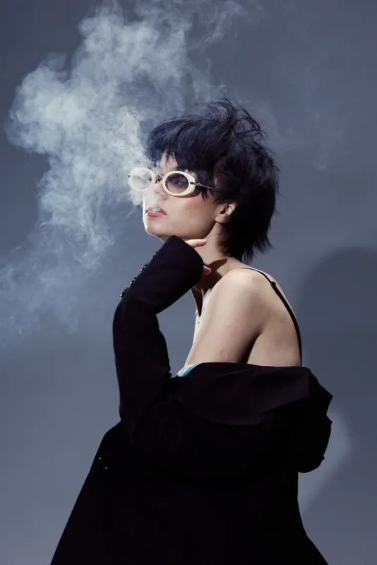 Stylish woman exhaling smoke with sunglasses wearing black clothing on dark background