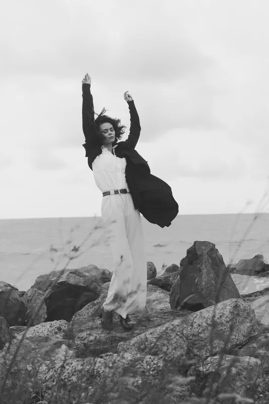 Monochrome image of a woman on a rocky shore with arms raised, flowing dress in the wind, ocean and sky behind, conveying freedom, movement and cinematic mood for fashion or lifestyle stock
