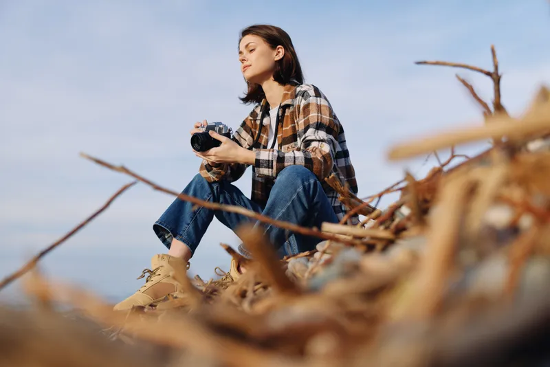 Woman photographer perched among driftwood, camera in hand, exploring coastal landscape for nature photography, outdoor exploration and focus on composition and mood.