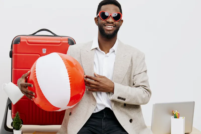 Smiling man with dark skin, sunglasses, beige blazer, white shirt, holding a beach ball, ready for vacation, in front of travel luggage, studio shot, lifestyle concept.