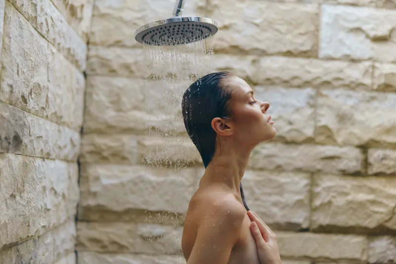 Woman enjoying relaxing outdoor shower under rainfall with closed eyes, natural stone wall background, wet hair, and serene expression, wellness and self-care concept.