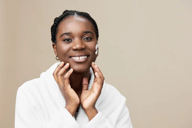 Happy young African woman in a white robe showcasing skincare routine with glowing skin, smiling in a minimalistic beige background