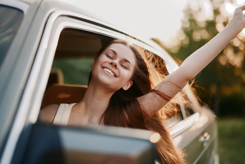 Happy young woman enjoying a road trip with her hair flowing in the wind, vibrant nature background, summer vibes, freedom and adventure concept.
