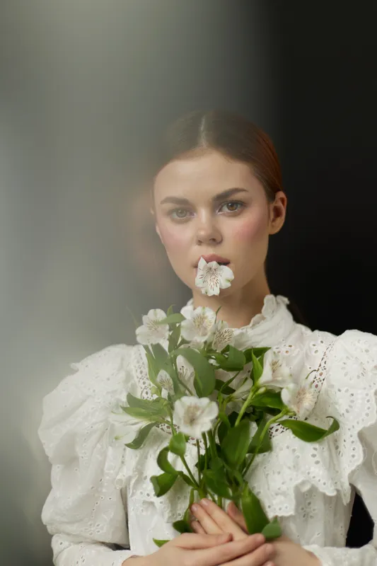 Young model in elegant white dress holding flowers and posing softly, radiating serenity and beauty with a delicate expression, ideal for spring themes