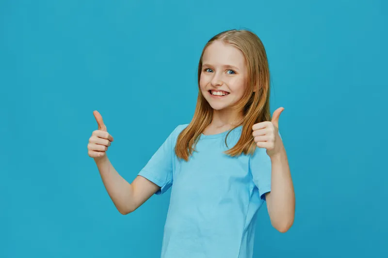 Happy girl giving thumbs up on blue background, portraying positivity and joy, wearing a light blue shirt, ideal for children s concepts and emotions