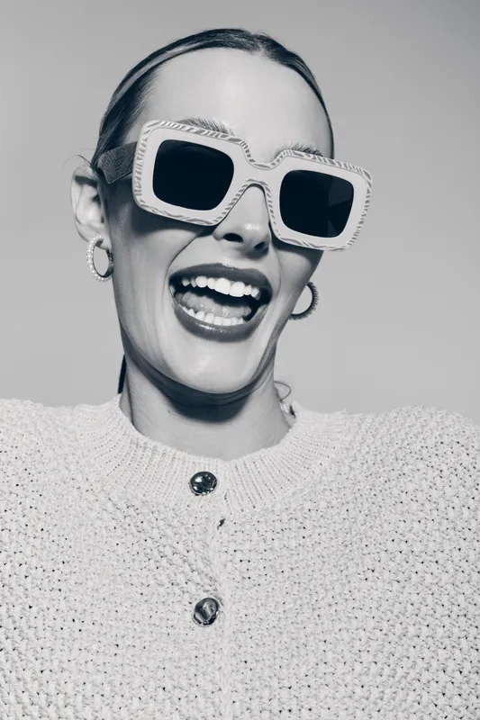 Smiling white woman with fair skin and slicked back hair wears large square sunglasses and knitted sweater. Studio portrait with joyful expression and fashion accessories concept.