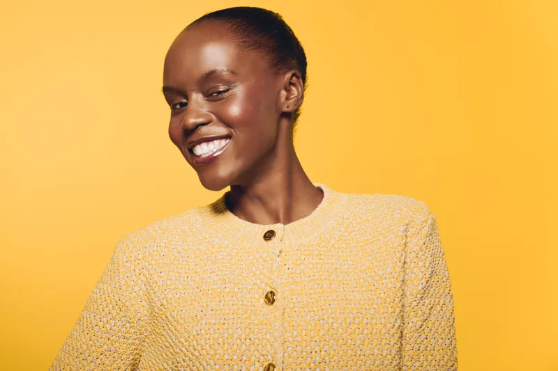 Smiling African woman with dark skin wearing a yellow sweater poses against a bright yellow background. Concept of happiness, beauty, confidence, and fashion portrait.