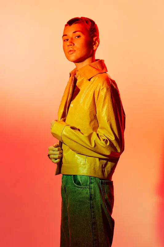 Stylish woman in yellow jacket posed against warm gradient background, showcasing modern fashion with a confident and bold expression in soft studio lighting.