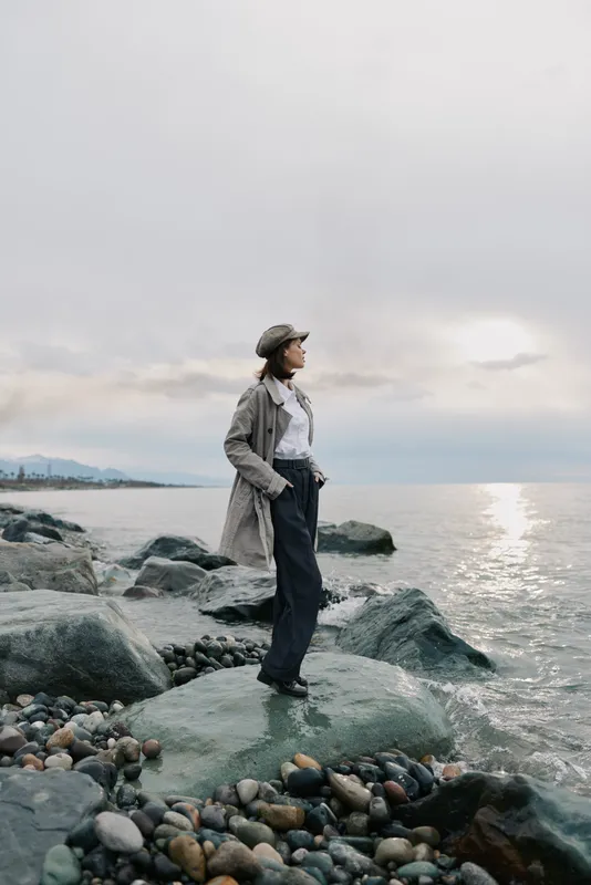 Woman stands on rocky shore wearing casual clothes and hat looking at calm sea under cloudy sky. Concept of contemplation, nature, travel, and peaceful outdoor moment by water.