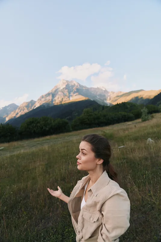 A young woman in casual beige jacket stands in a grassy field with mountains in the background under a clear sky. Lifestyle concept of nature exploration and outdoor relaxation.
