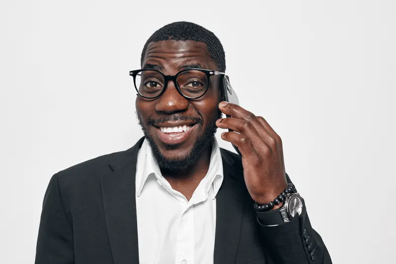 African American man with dark skin wearing glasses and black suit smiling while talking on smartphone against white background. Confident businessman in formal outfit with wristwatch and beard.