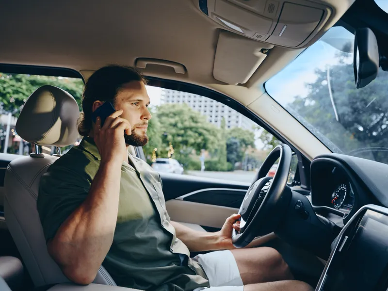 A man sits in a car, driving with one hand on the wheel while talking on a mobile phone, focused expression, daytime street scene visible through the window.