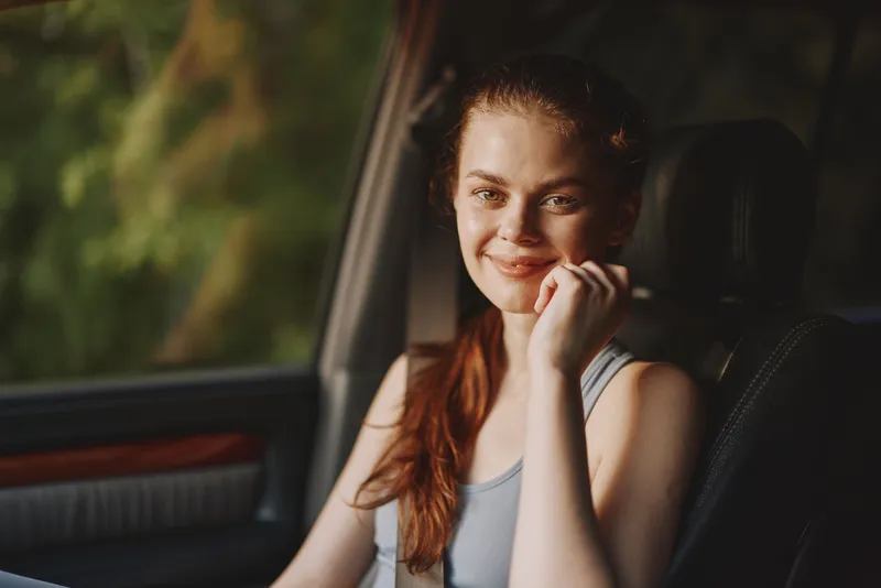 Smiling young woman with long hair, wearing a casual tank top, sitting in a car, surrounded by nature, bright sunlight, summer vibes, urban lifestyle