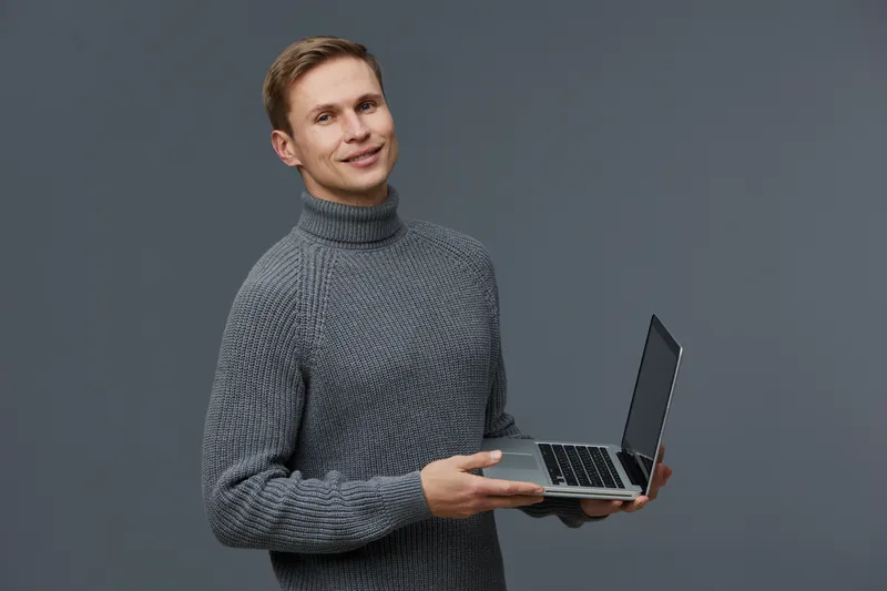 Smiling man holding a laptop in hands wearing a gray knitted turtleneck sweater isolated on plain gray background studio portrait. People lifestyle concept