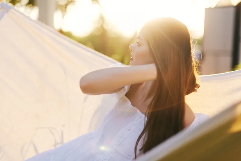 Young woman relaxing in a hammock during sunset, wearing a white dress, exuding tranquility and serenity in a beautiful outdoor setting