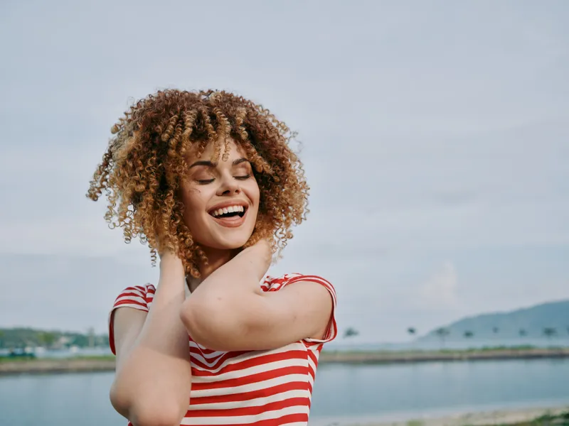 Joyful portrait of a young woman with curly hair and a striped shirt, standing beside water, smiling with eyes closed and arms raised to touch her neck.
