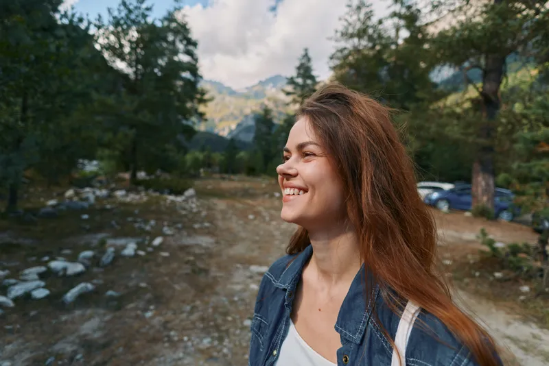 A cheerful woman stands outdoors in a mountain setting, smiling warmly as she gazes toward the distance, exuding happiness and freedom in nature's expansive scenery.