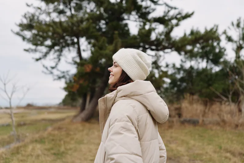 Outdoor scene with a person wearing a beige winter coat and knit beanie, standing in a park among trees and open field, calm weather and reflective mood.