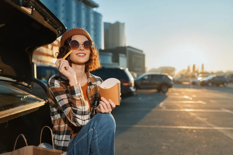 woman eating fries by car in a parking lot at golden hour, smiling with sunglasses and beanie, candid lifestyle scene of authenticity, mindful living and emotional storytelling.