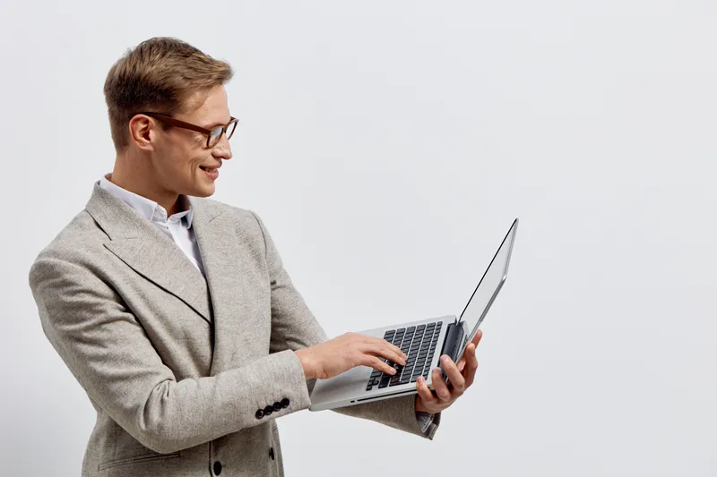 Professional happy young Caucasian man in glasses wearing beige blazer and white shirt using laptop, smiling and looking at screen, isolated on plain white background in studio portrait. Workplace lif