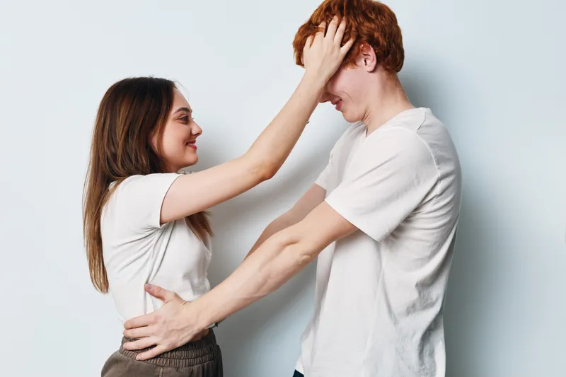 Young couple interacting with affection and playfulness, woman touching man head with hands, smiling and showing connection in casual clothing on white background.