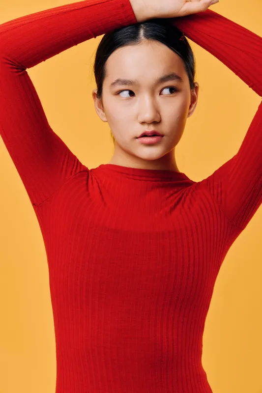 Young woman in red sweater posing with arms raised against yellow background. Portrait of fashionable female model with natural look and black hair pulled back for casual style.