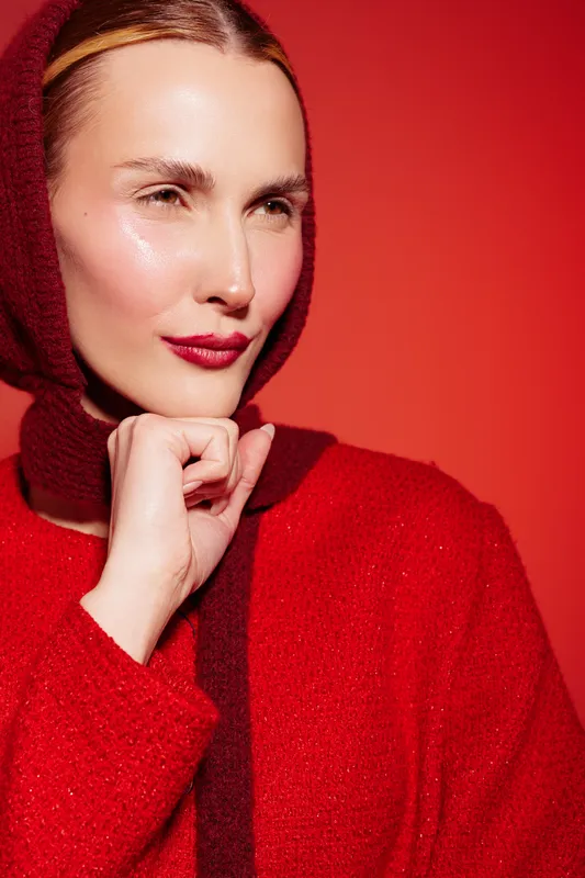 Portrait of a woman in red sweater and headscarf posing against red background. She has glossy skin and red lipstick, creating a bold and stylish fashion concept with warm lighting.