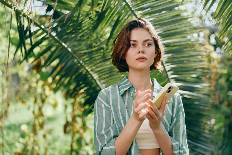 Portrait of a woman standing among lush palm leaves, holding a smartphone, with soft natural light and a relaxed, stylish mood in an outdoor tropical setting.
