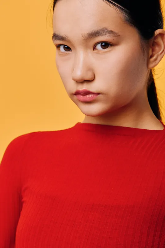Portrait of young woman with natural makeup and serious expression wearing red sweater on yellow background. Closeup of female model with sleek hair and smooth skin for beauty and fashion.
