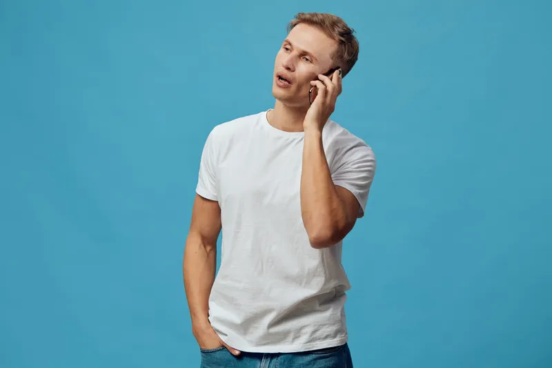 Man talking on smartphone isolated on blue background. Casual young male model with blonde hair in white t-shirt and jeans engaging in conversation. Studio portrait with space for advertising or comme