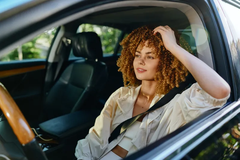 Young woman with curly hair driving car, wearing casual light jacket, looking relaxed and confident in bright natural light.