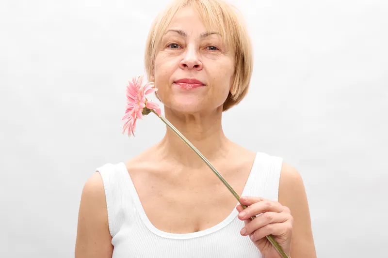 Woman flower spring smile beauty wellness skincare aging portrait studio, elderly woman holding pink flower on a stick, white background, soft light, calm happiness, wellbeing