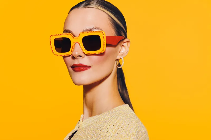 Stylish woman with slicked hair wears oversized orange sunglasses and red lipstick against bright yellow background. Concept of fashion, summer, and confident beauty portrait.